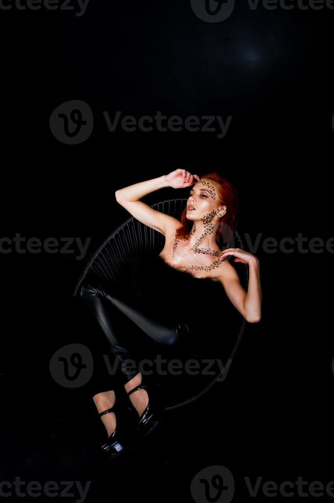 Fashion model red haired girl with originally make up like leopard predator isolated on black. Studio portrait on chair. photo