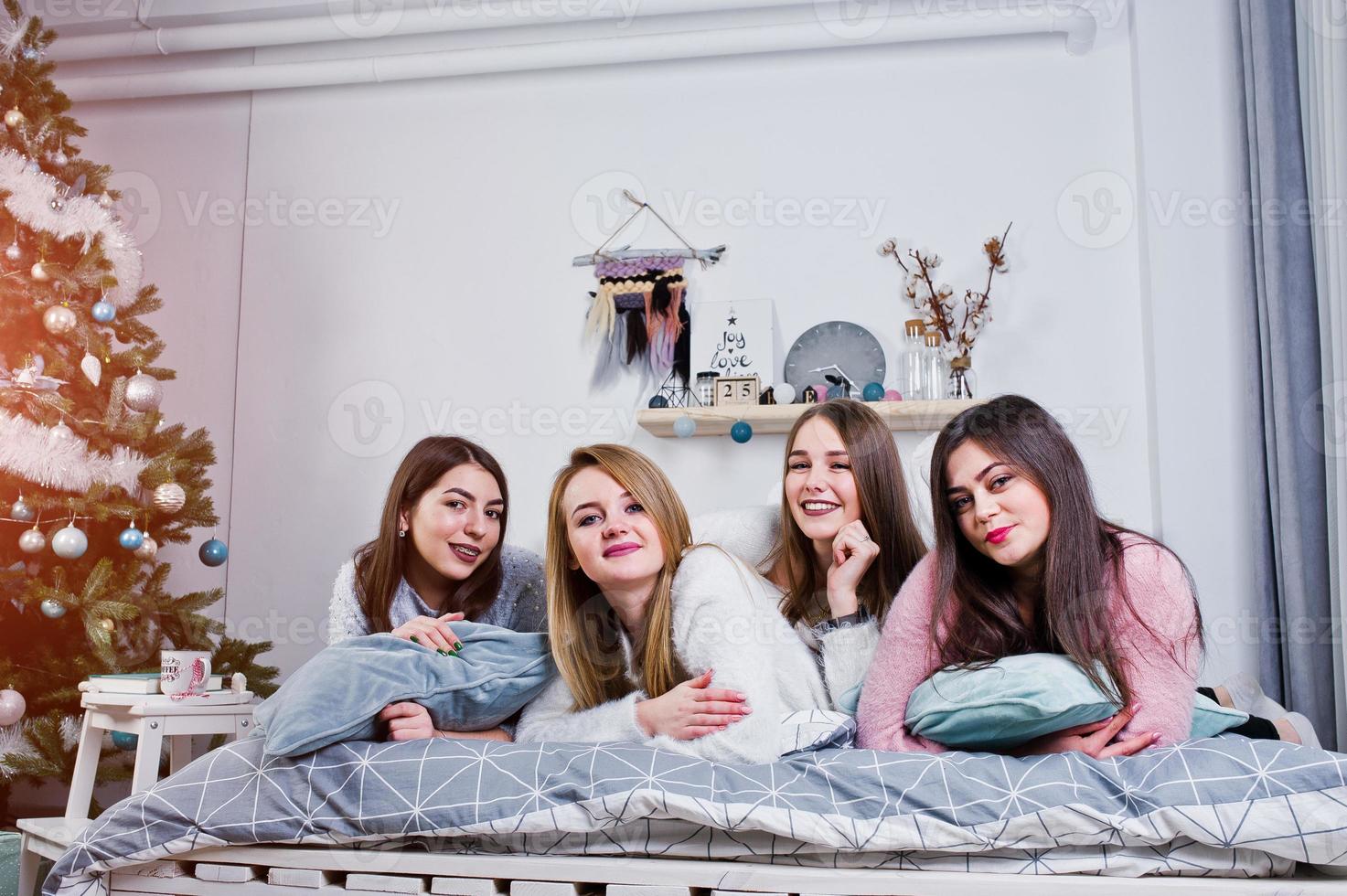 Four cute friends girls wear on warm sweaters and black pants on bed at new year decorated room on studio, play with pillows. photo
