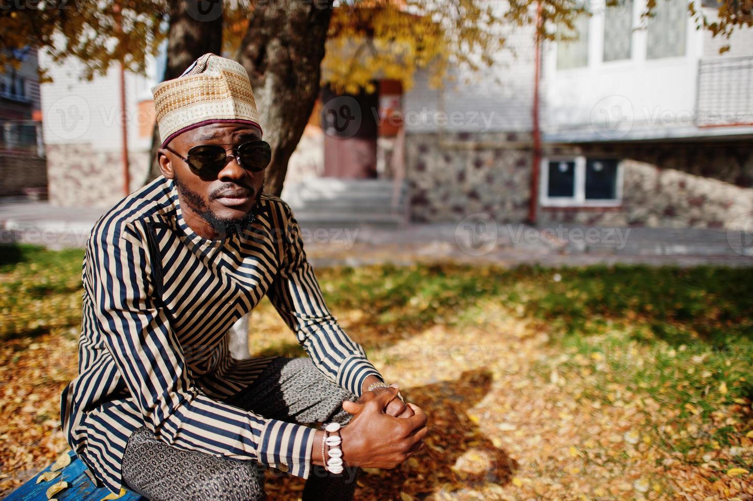 Portrait of stylish black african american man at hat and sunglasses against sunny autumn fall background. Rich people in africa at traditional dress. photo