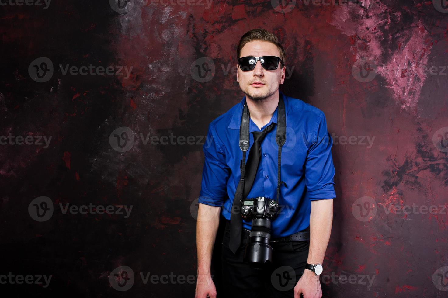 Studio portrait of stylish professional photographer man with camera, wear on blue shirt and necktie, sunglasses. photo