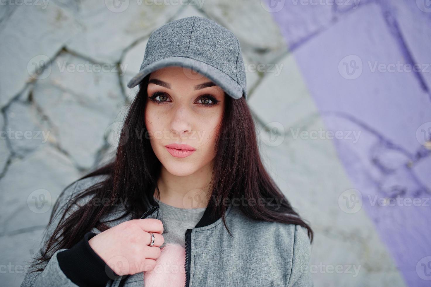 Stylish brunette girl in gray cap, casual street style on winter day against colored wall. photo