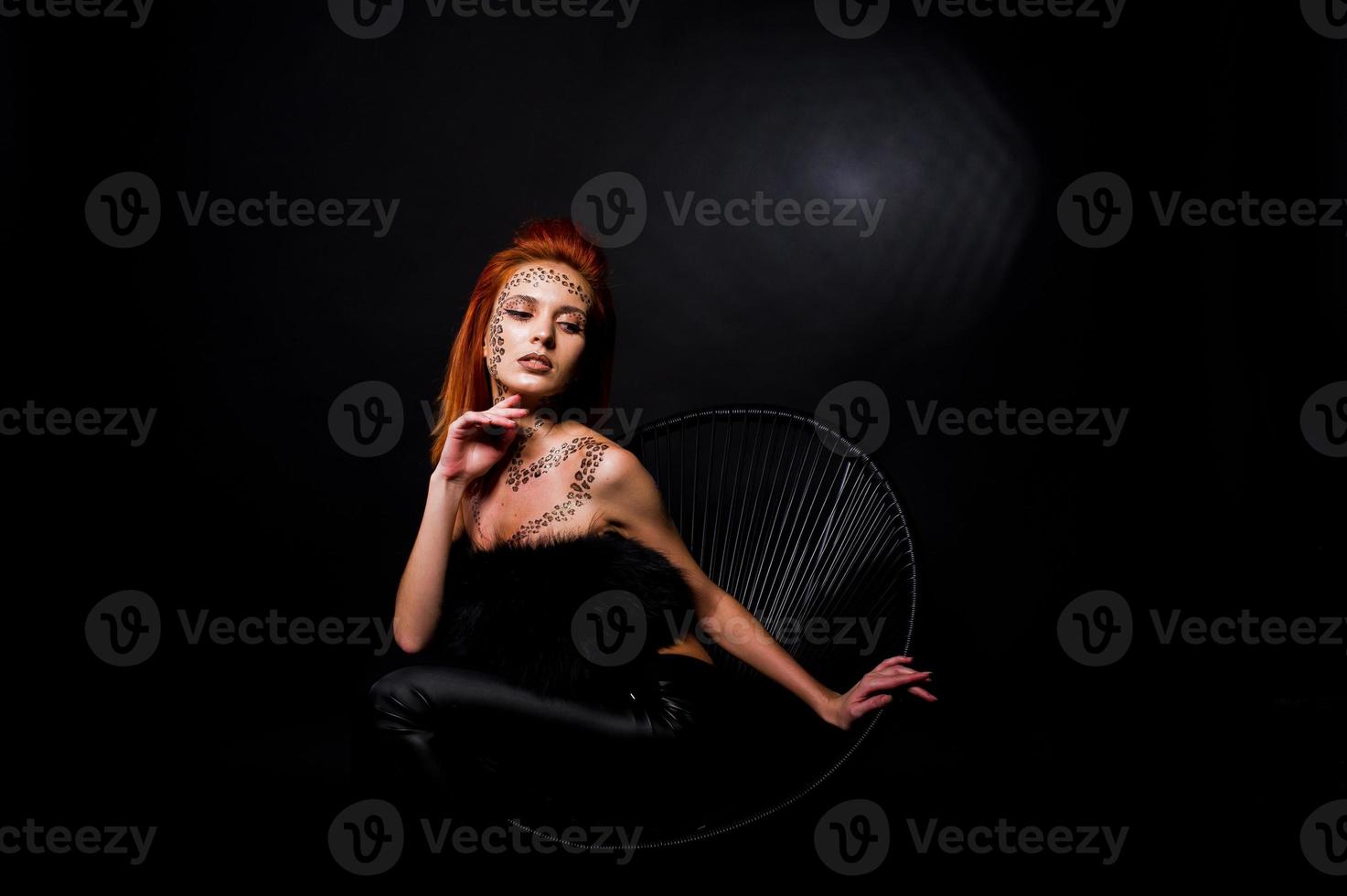 Fashion model red haired girl with originally make up like leopard predator isolated on black. Studio portrait on chair. photo