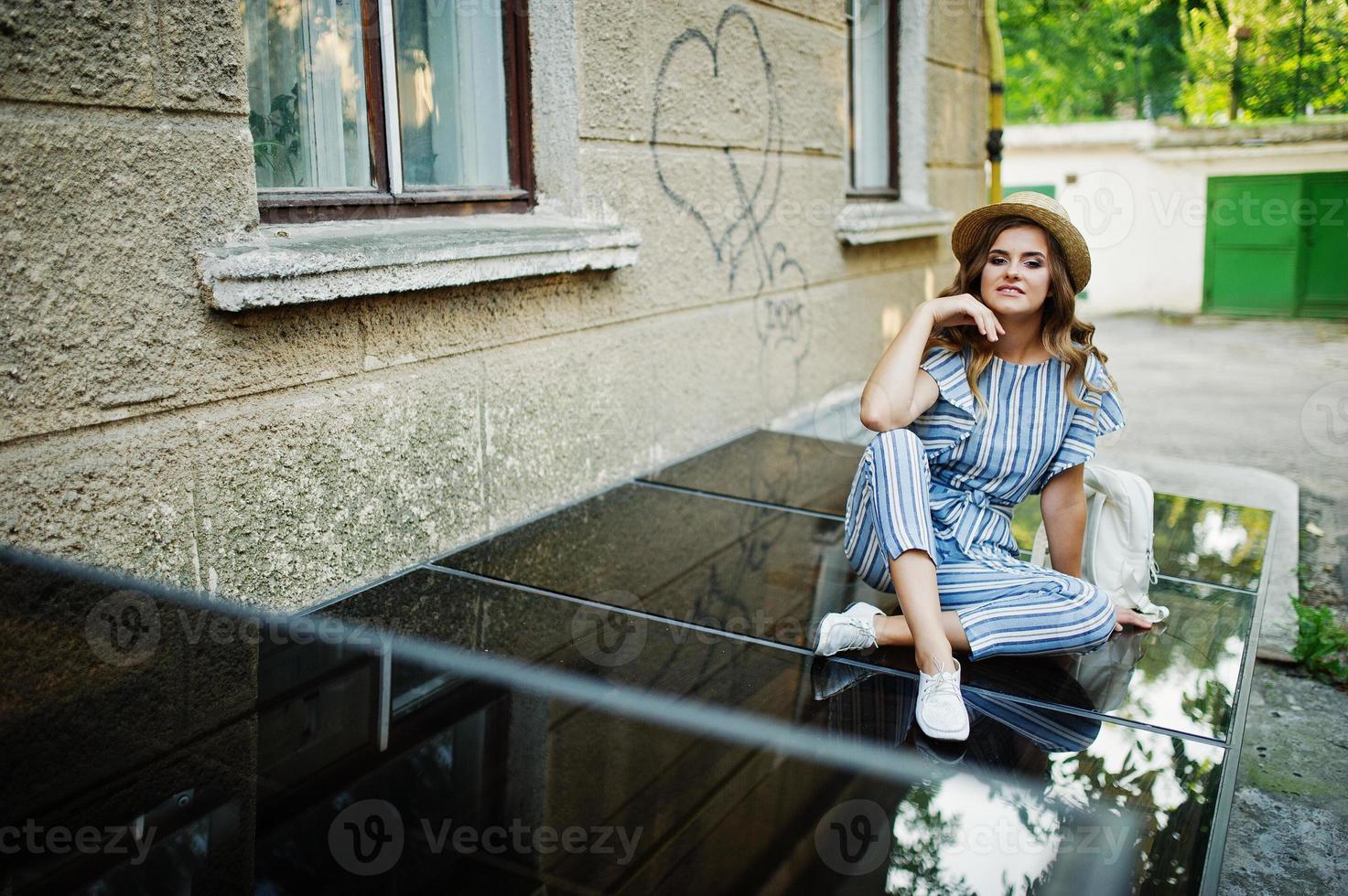 Portrait of a fabulous young woman wearing striped overall and hat sitting on black shiny surface with backpack next to the building. photo
