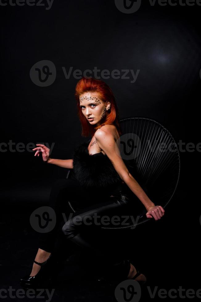 Fashion model red haired girl with originally make up like leopard predator isolated on black. Studio portrait on chair. photo