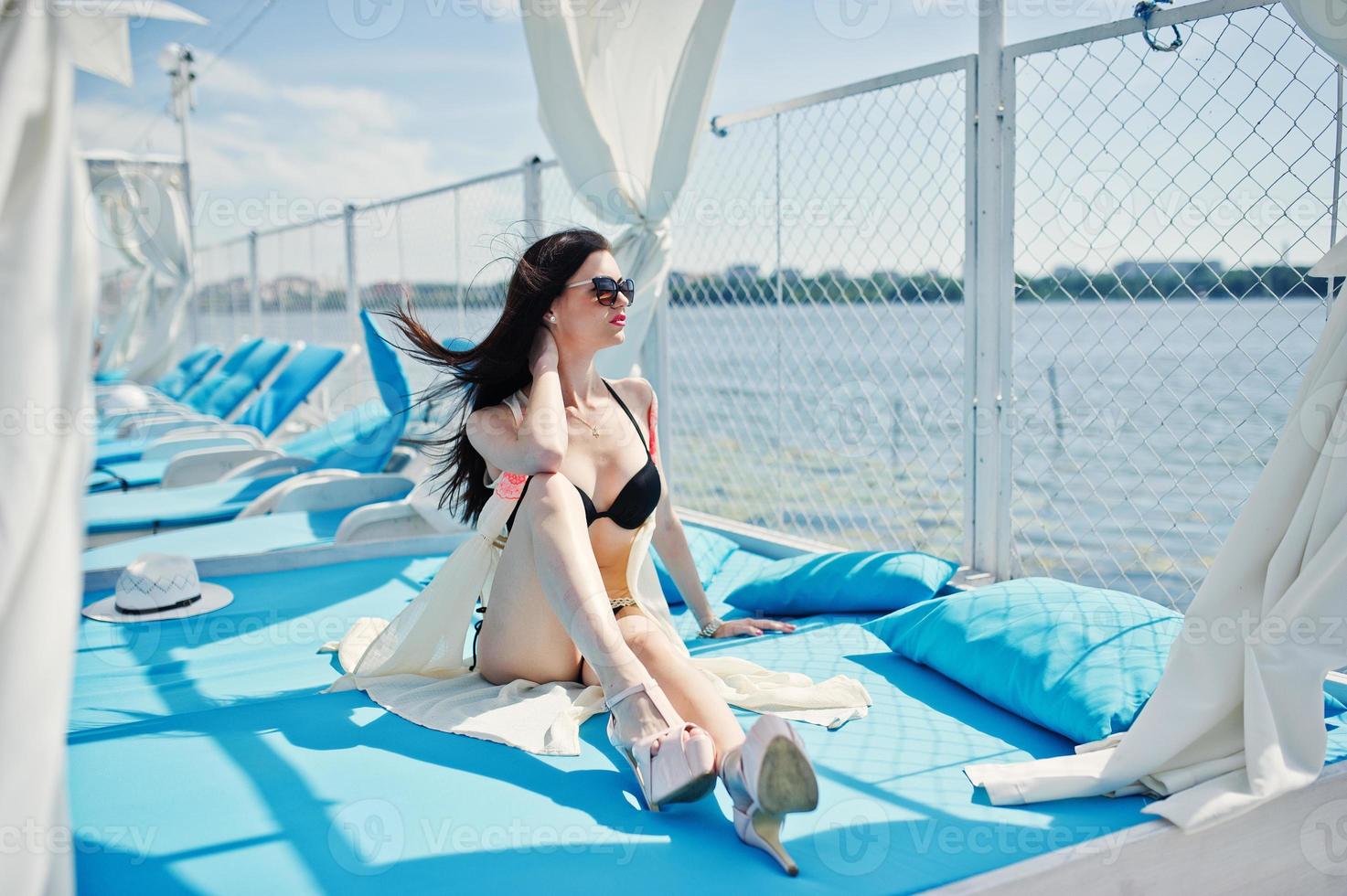 Portrait of an attractive brunette in black bikini sitting on the lounger with sunglasses on by the lake. photo
