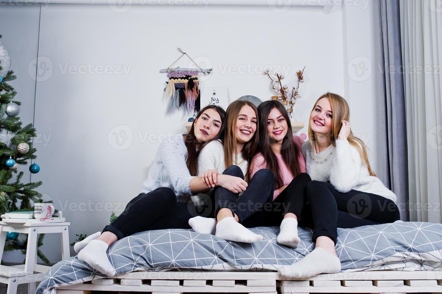 Four cute friends girls wear on warm sweaters and black pants on bed at new year decorated room on studio. photo