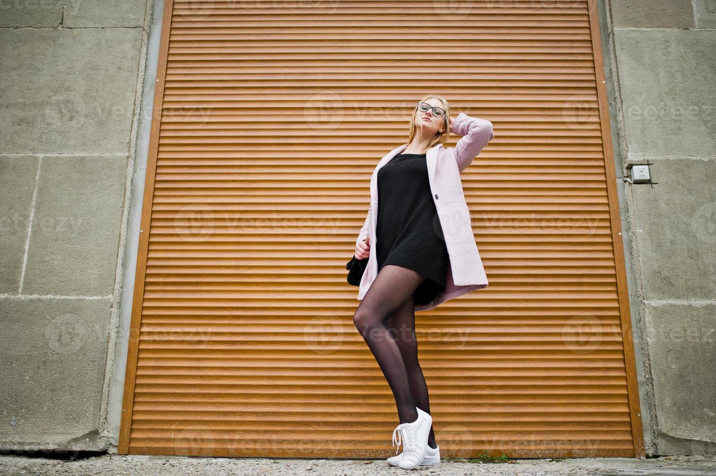 Blonde girl at glasses and pink coat against shutters. photo