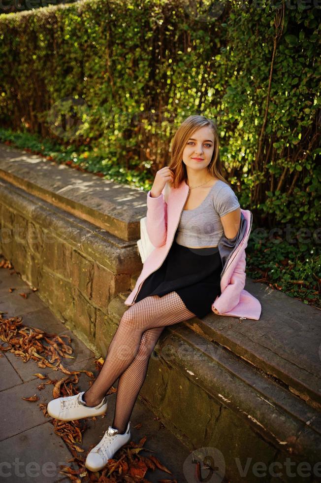 Young blonde girl in black skirt and pink coat posed at sunny day on street. photo