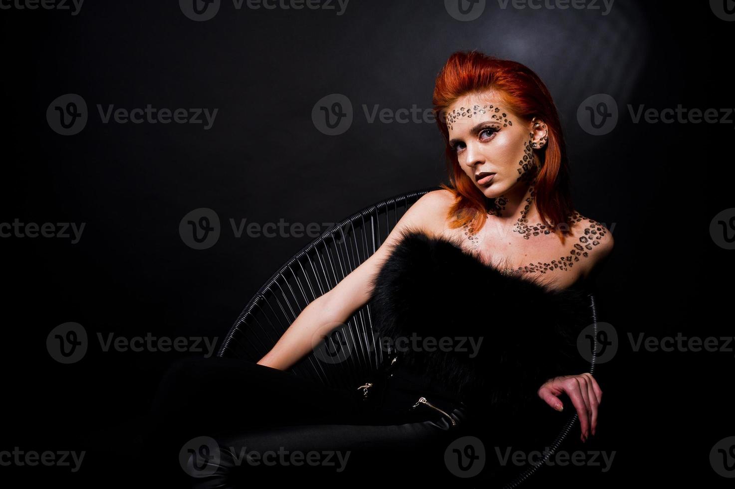 Fashion model red haired girl with originally make up like leopard predator isolated on black. Studio portrait on chair. photo