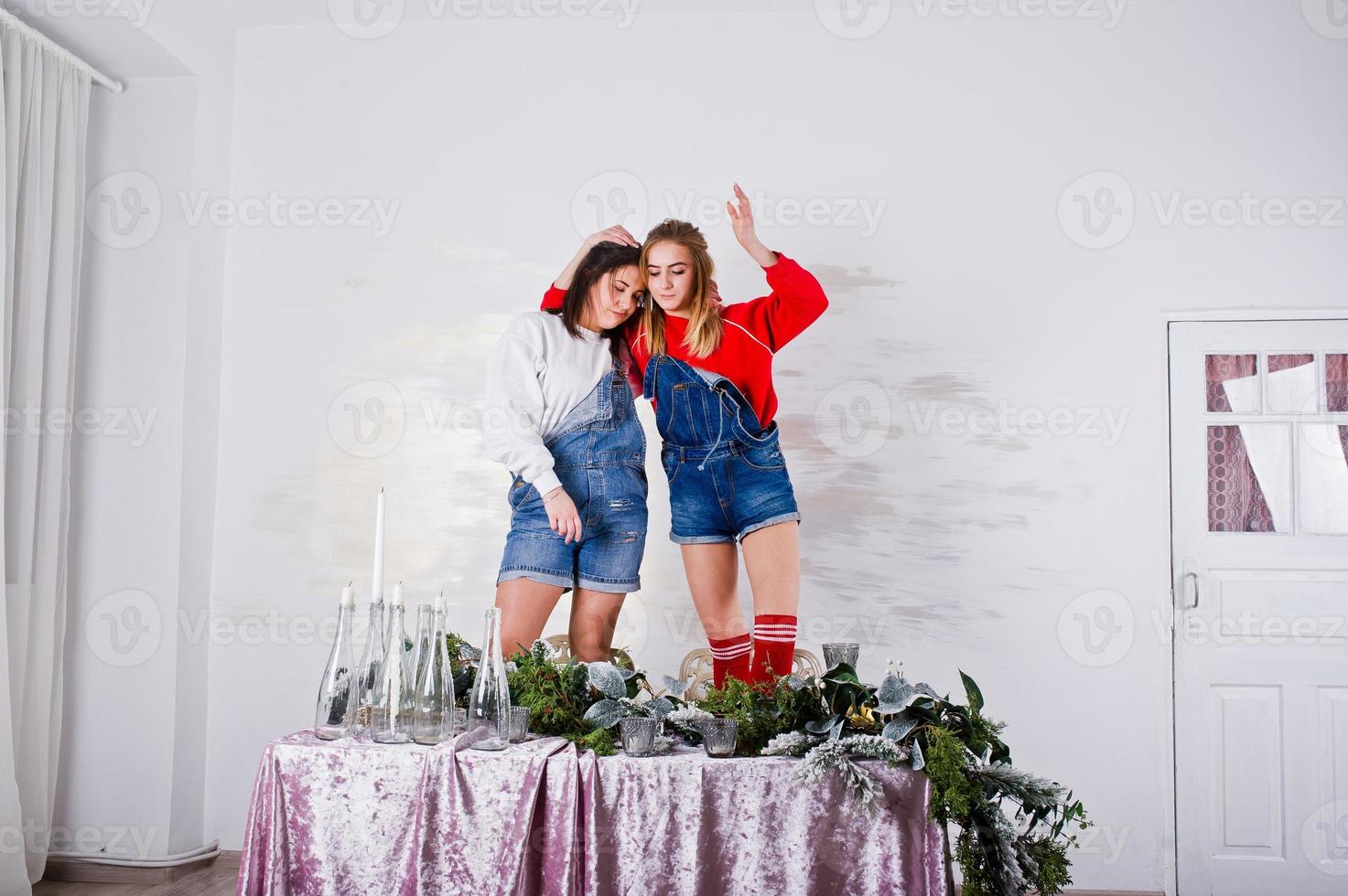 dos amigas hermosas y divertidas visten pantalones cortos de jeans y polainas contra la mesa con decoración de año nuevo. foto