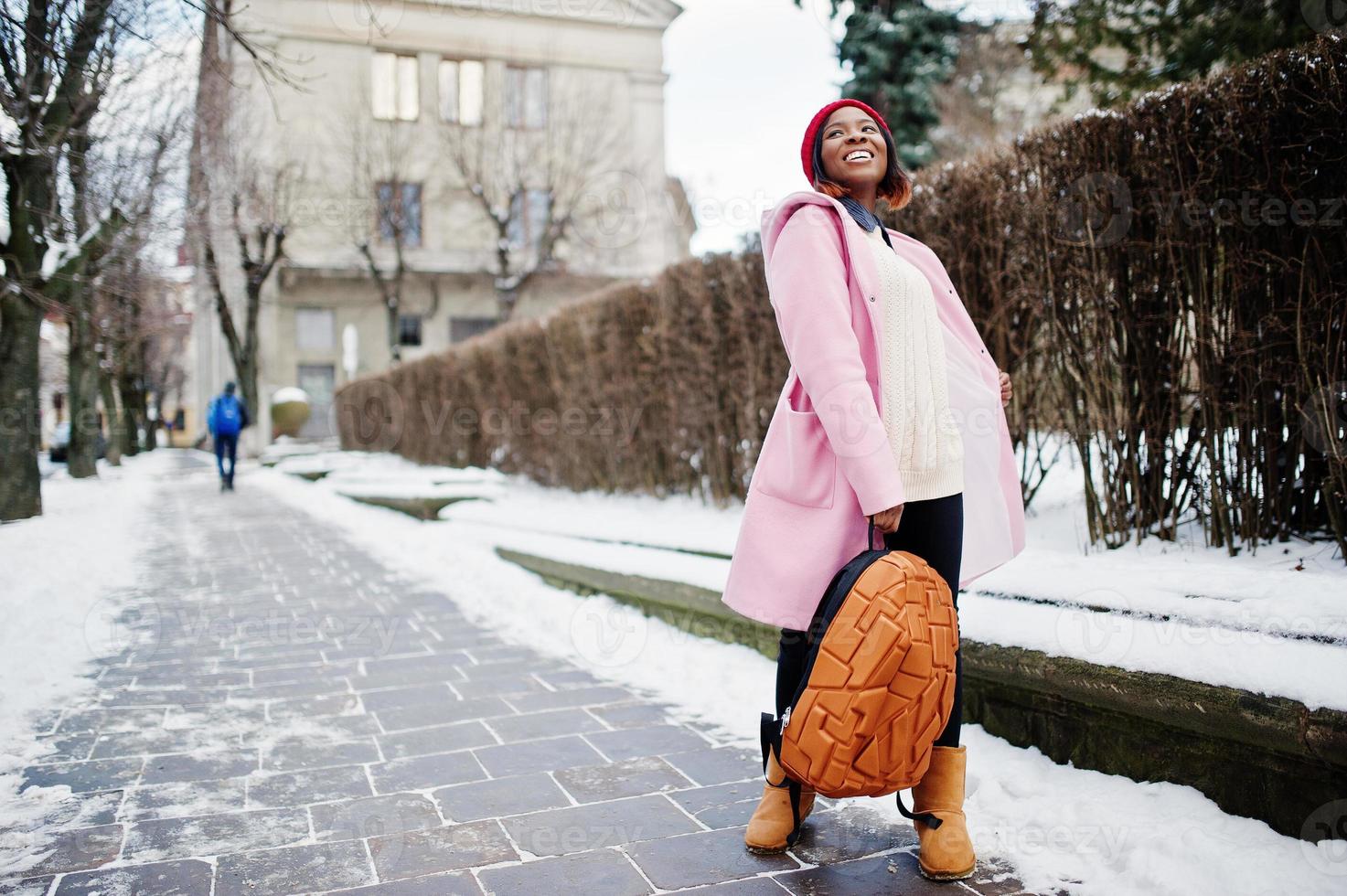 niña afroamericana con sombrero rojo y abrigo rosa con mochila en la calle de la ciudad el día de invierno. foto