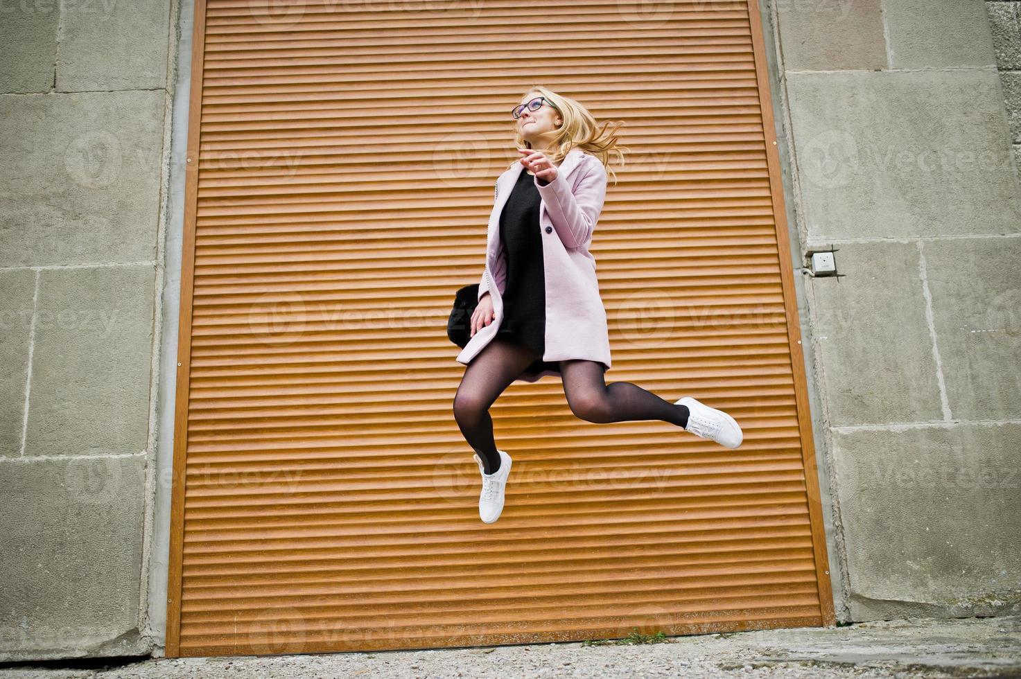 Blonde girl at glasses and pink coat with handbag jump against shutters. photo
