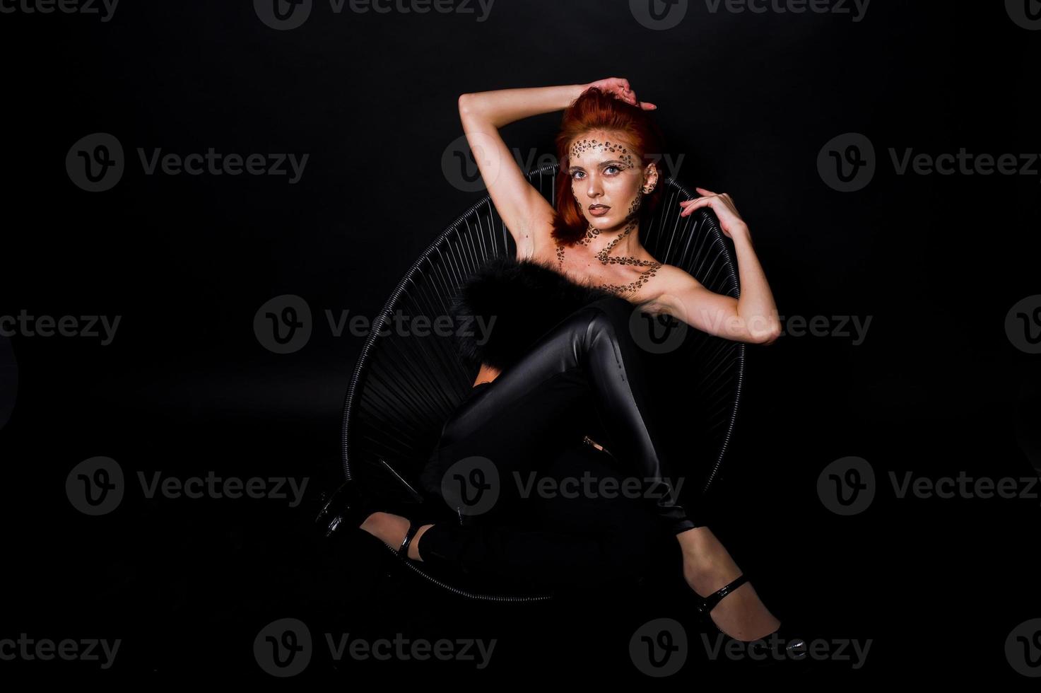 Fashion model red haired girl with originally make up like leopard predator isolated on black. Studio portrait on chair. photo