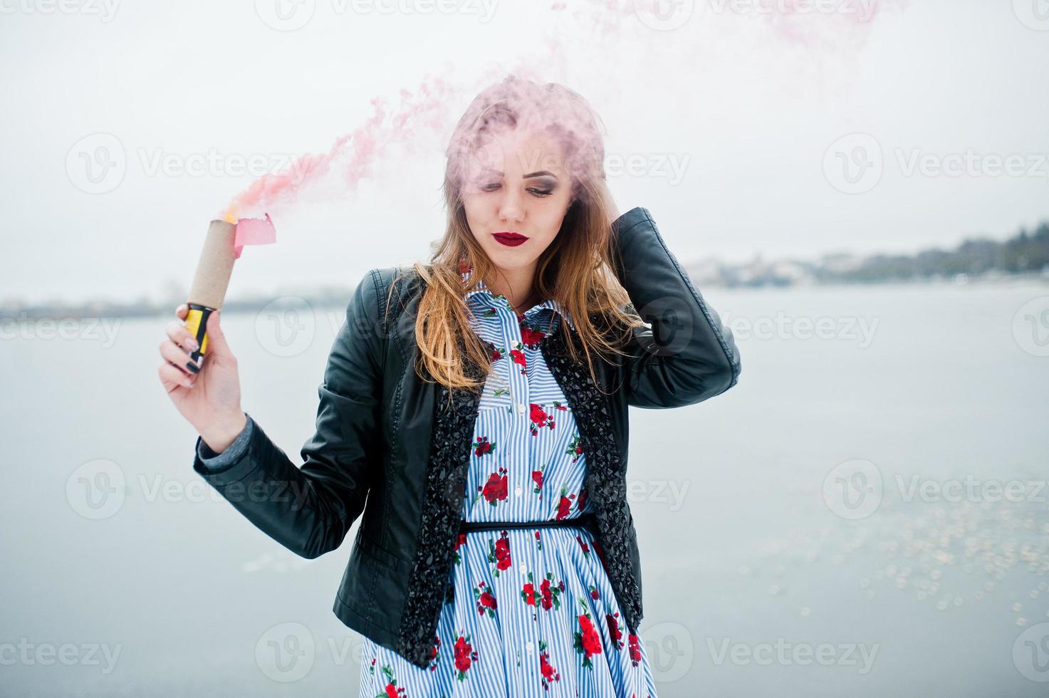 chica elegante con chaqueta de cuero sostiene una bengala de humo rosa en el día de invierno contra el lago congelado. foto