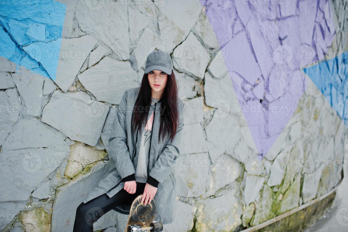 Stylish brunette girl in gray cap, casual street style with skate board on winter day against colored wall. photo