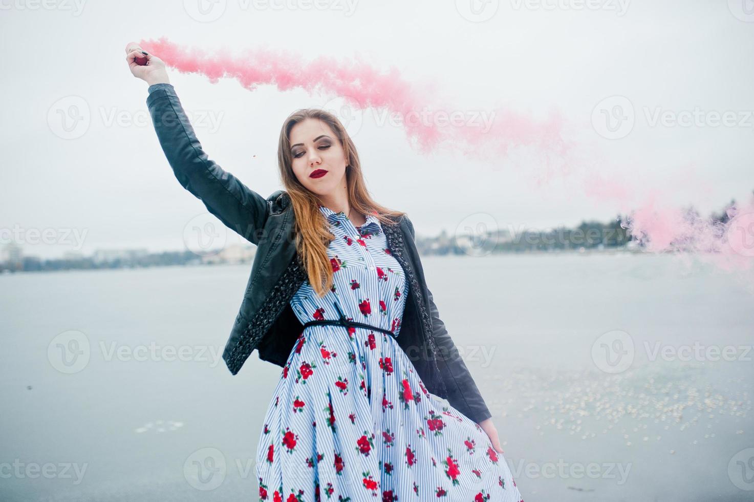 chica elegante con chaqueta de cuero sostiene una bengala de humo rosa en el día de invierno contra el lago congelado. foto