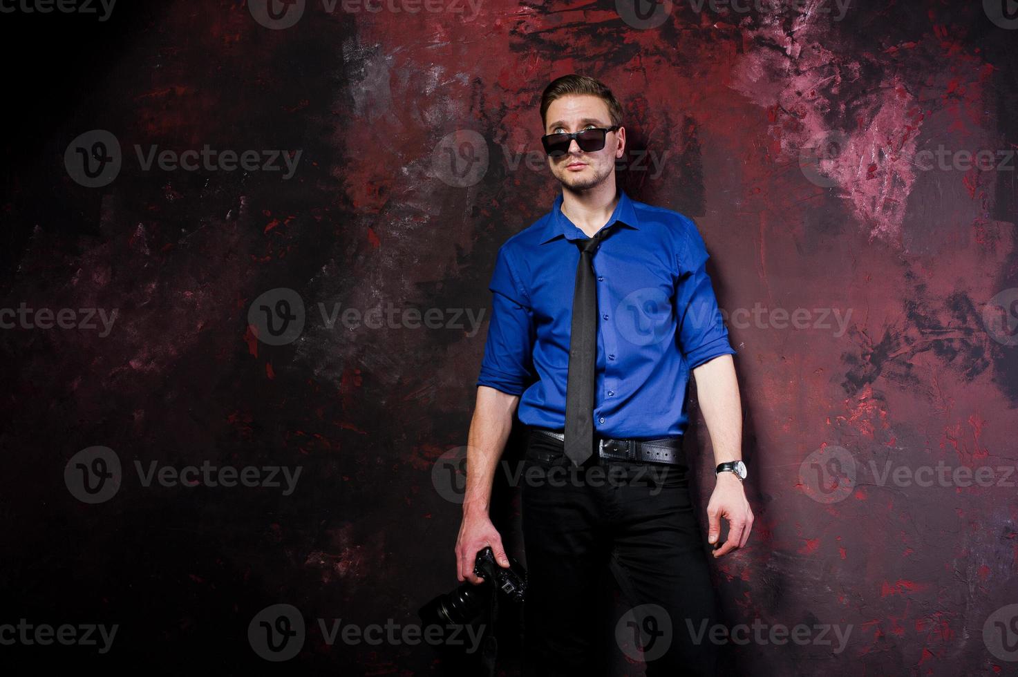 Studio portrait of stylish professional photographer man with camera, wear on blue shirt and necktie, sunglasses. photo
