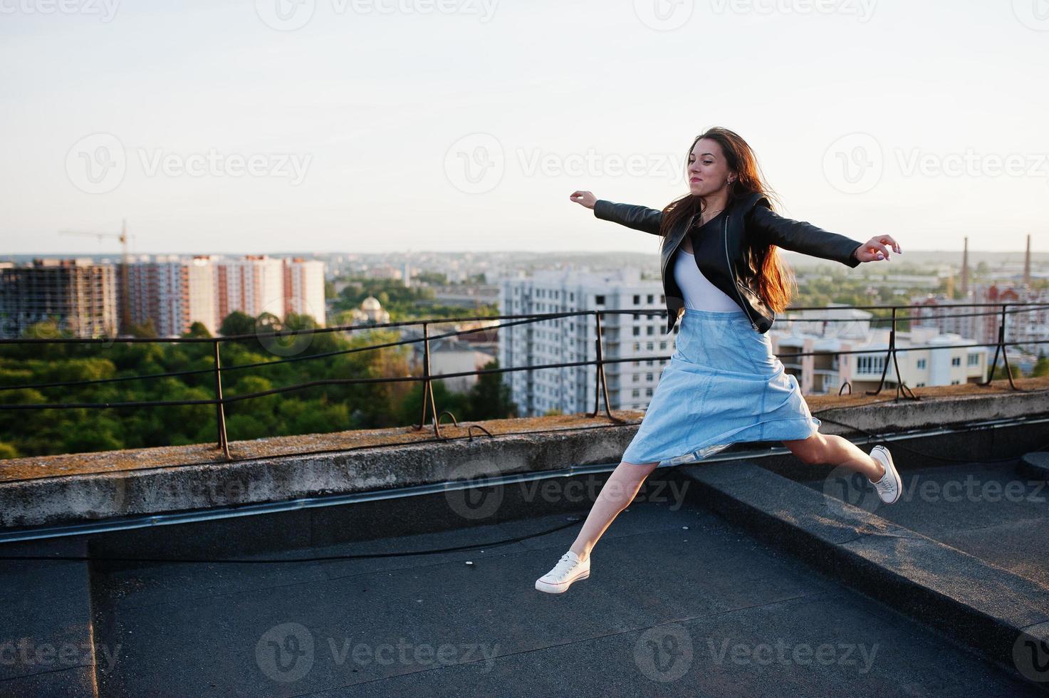 retrato de una bella joven con chaqueta de cuero negro y vestido azul saltando en un tejado. foto