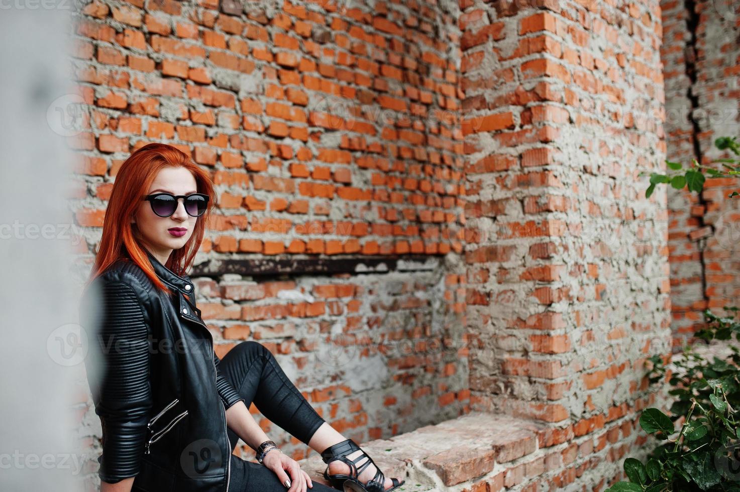 Red haired stylish girl in sunglasses wear in black, against abadoned place with brick walls. photo