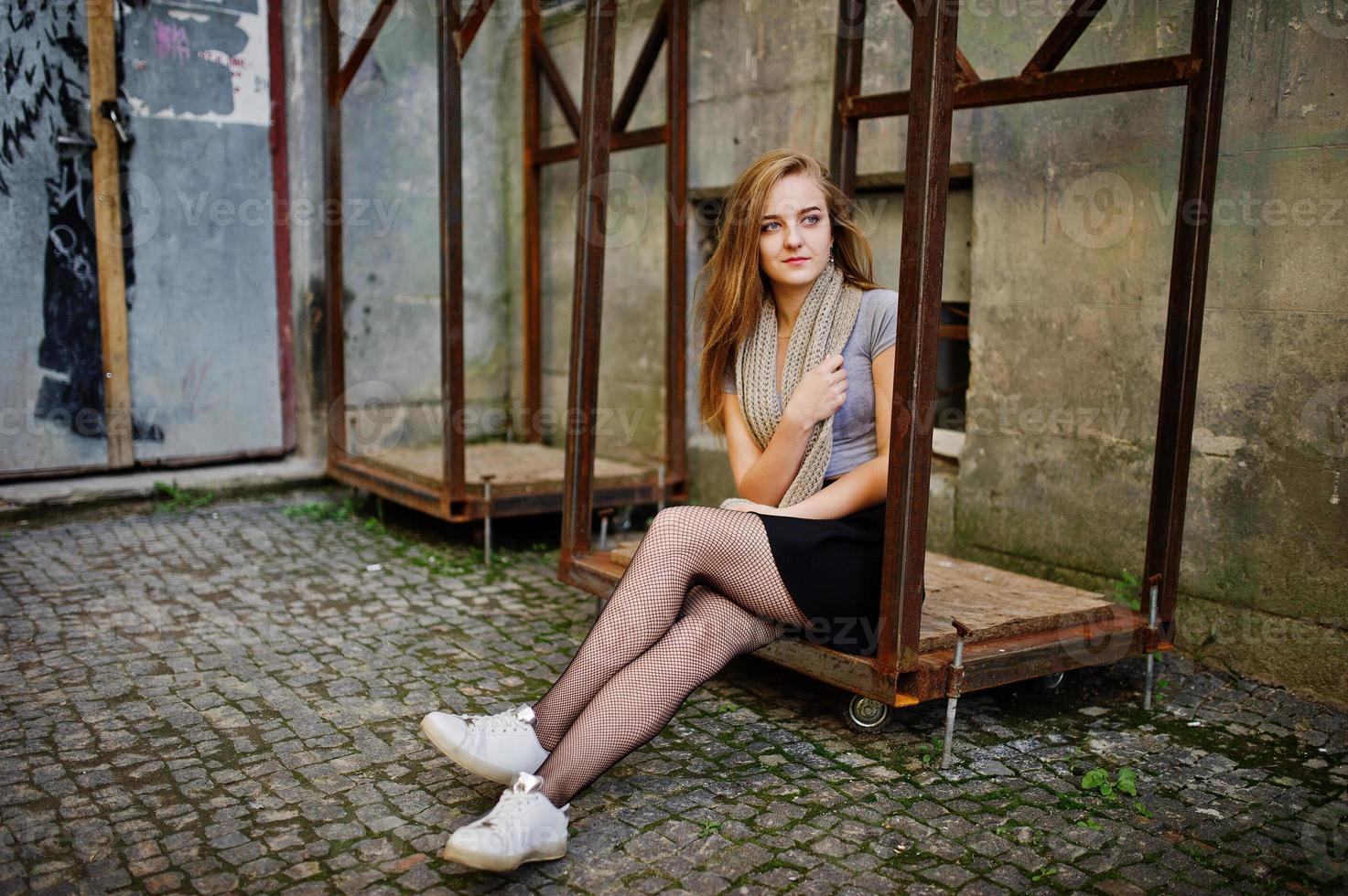 Young blonde girl in black skirt with scarf posed at street of city near the iron armor. photo