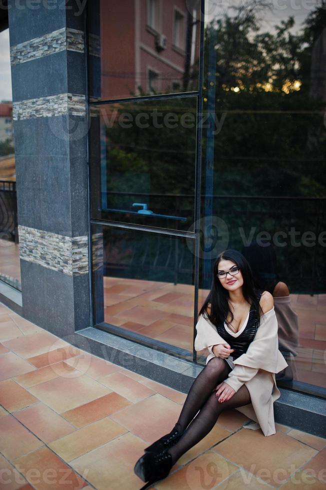 mujer sexy de pelo negro con gafas y abrigo posado contra un edificio con ventanas modernas. foto