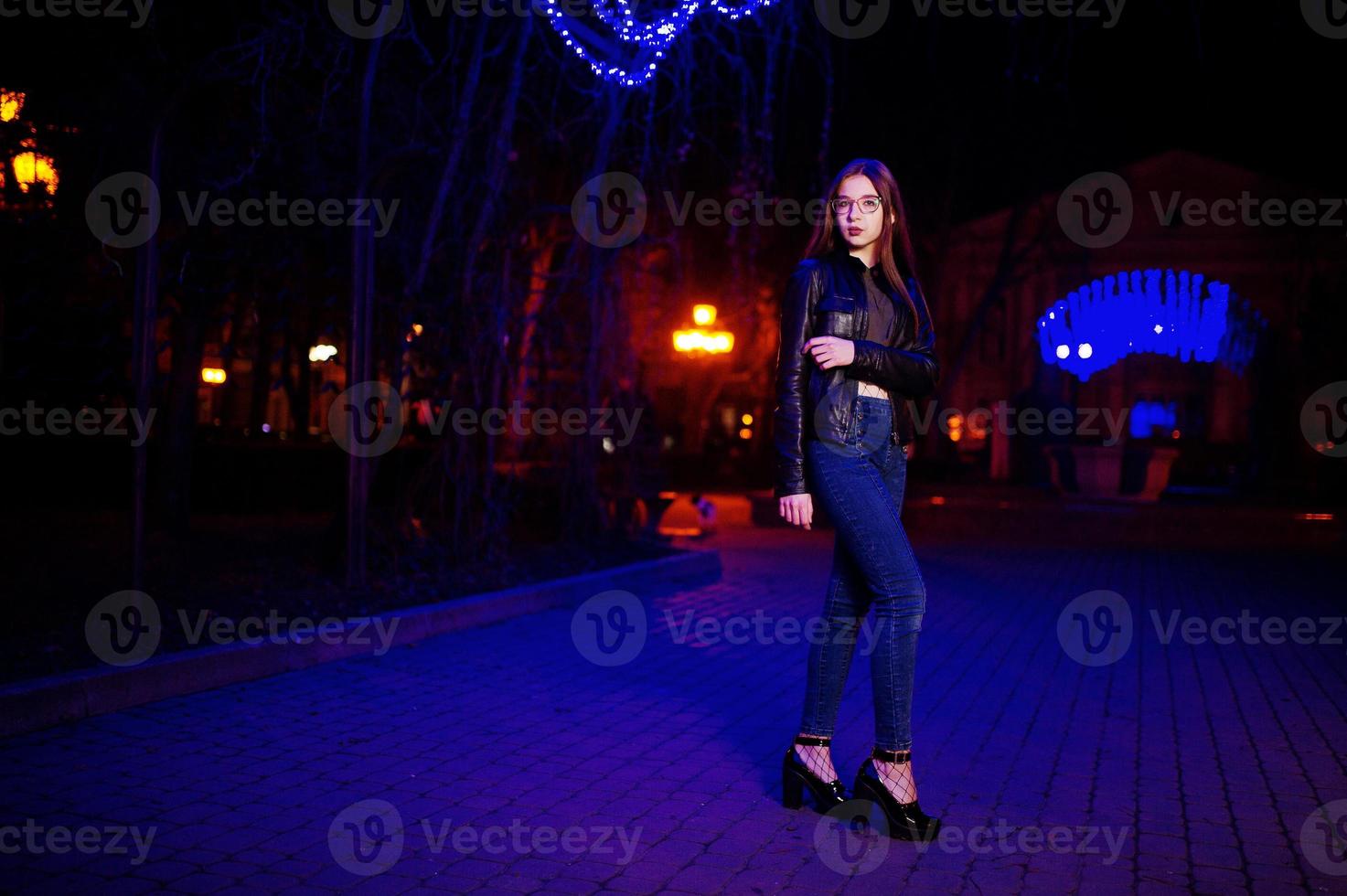 Night portrait of girl model wear on glasses, jeans and leather jacket against blue lights garland of city street. photo