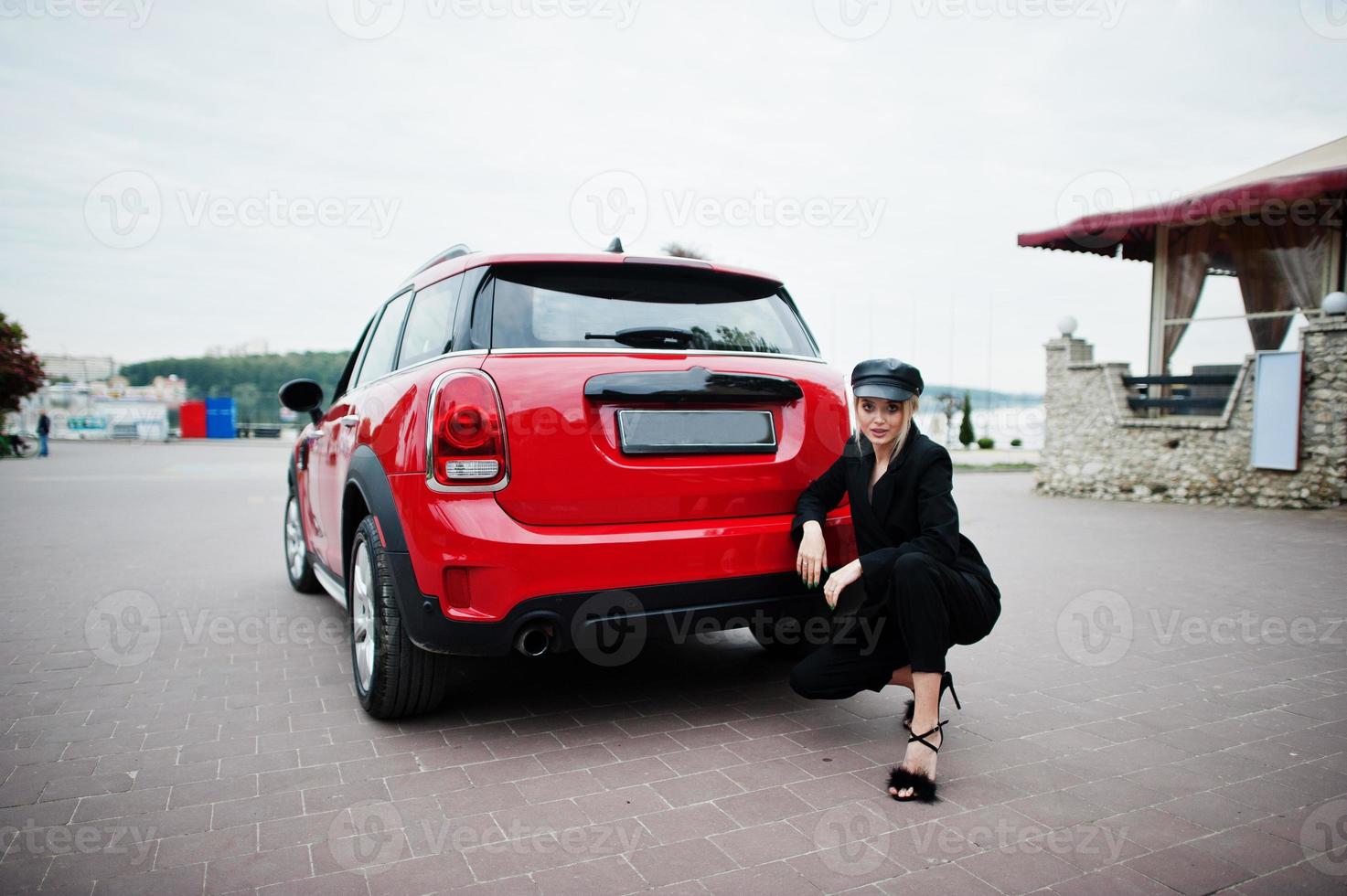 retrato de una hermosa modelo de mujer rubia sexy con gorra y todo negro con maquillaje brillante cerca del coche rojo de la ciudad. foto