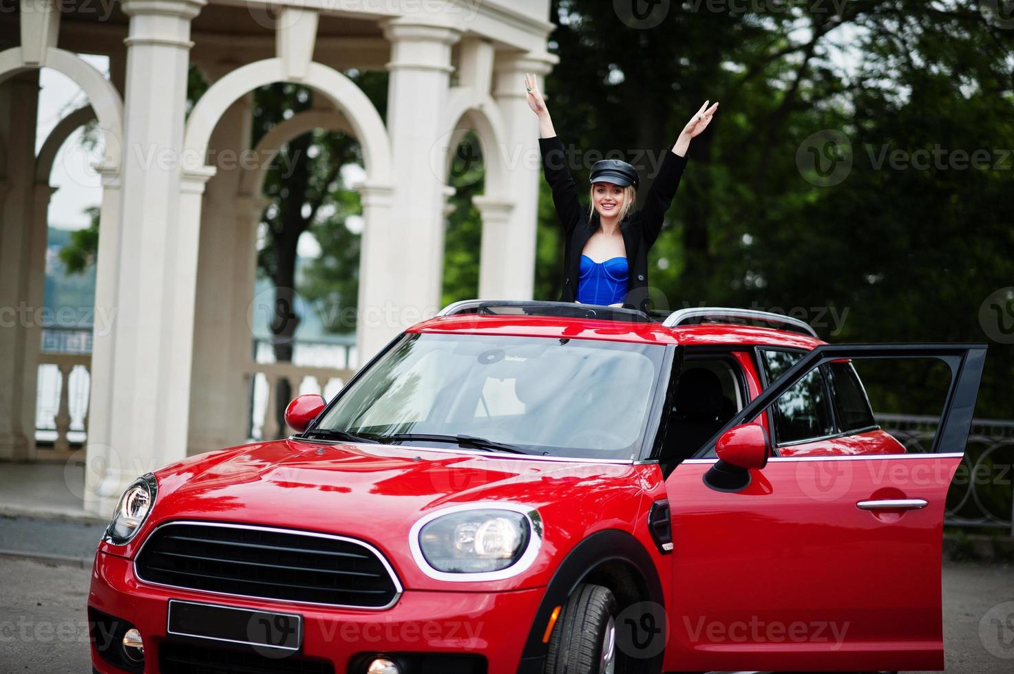 retrato de una hermosa modelo rubia de moda sexy con gorra y todo negro con maquillaje brillante en el techo solar abierto del coche rojo de la ciudad. foto