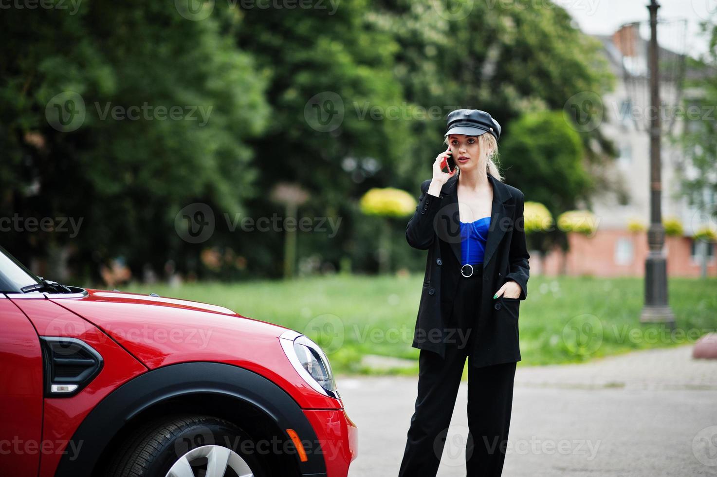 Portrait of beautiful blonde sexy fashion woman model in cap and in all black with bright makeup near red city car, with mobile phone. photo