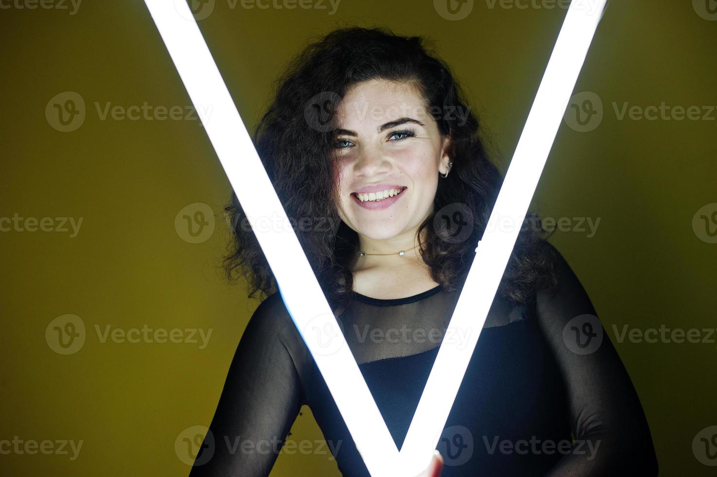 Curly brunette plus size model with two long led lamp against yellow wall. photo