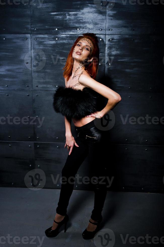 Fashion model red haired girl with originally make up like leopard predator against steel wall. Studio portrait. photo
