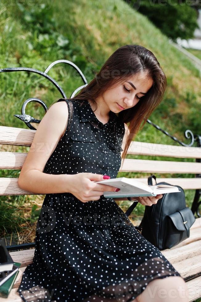 Portrait of a fabulous woman in black polka dot dress sitting on the bench and reading in the park. photo