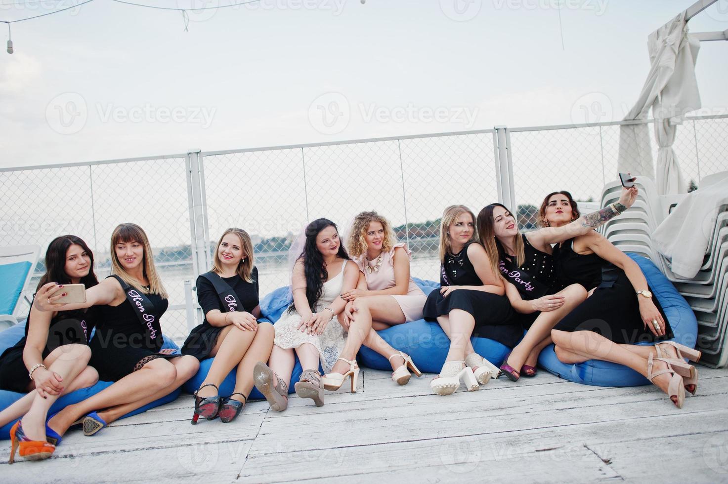 Group of 8 girls wear on black and 2 brides at hen party sitting on pillows and making selfie at pier on beach side. photo