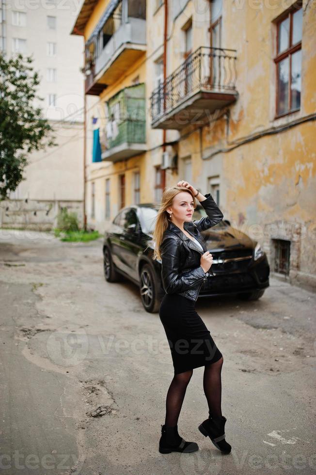 elegante chica rubia vestida con una chaqueta de cuero negro posando en las calles del coche de lujo de fondo de la ciudad. foto