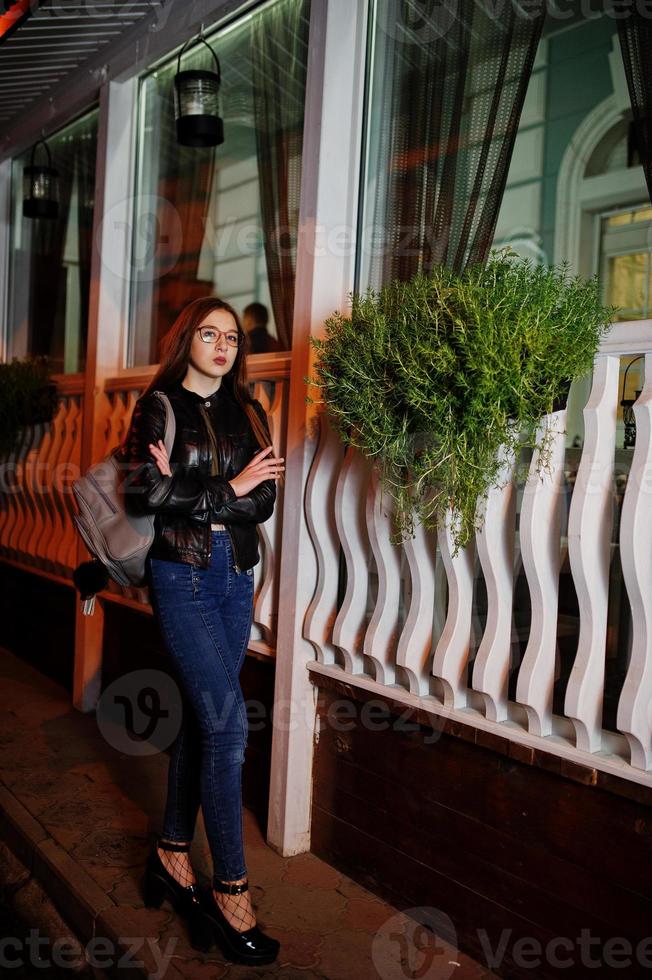 retrato nocturno de modelo de niña con gafas, jeans y chaqueta de cuero, con mochila, contra las calles de la ciudad. foto