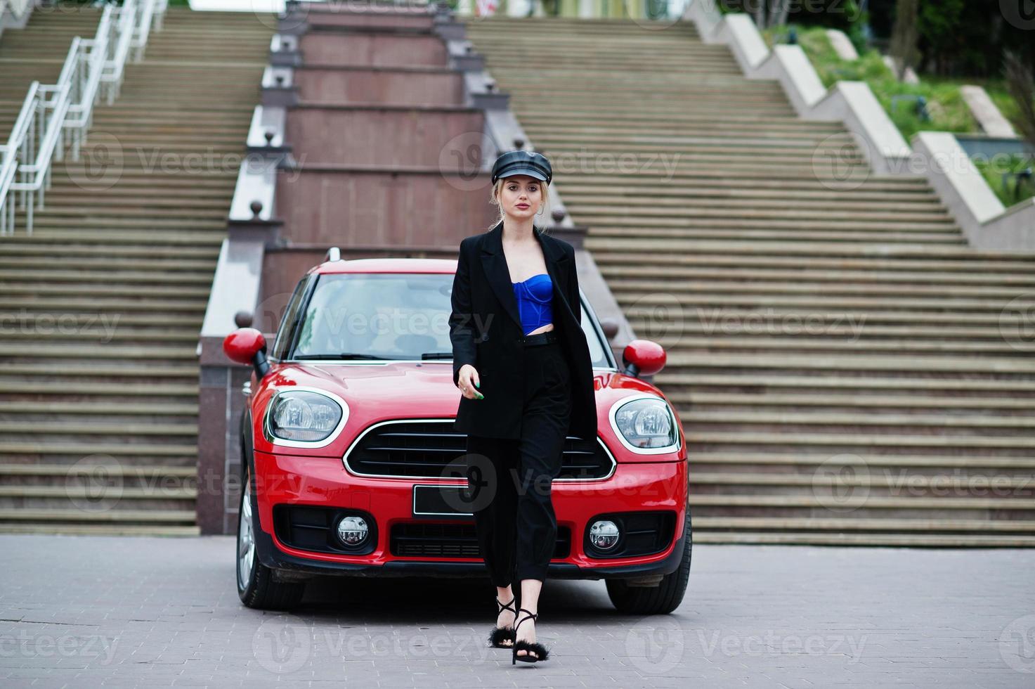 Portrait of beautiful blonde sexy fashion woman model in cap and in all black, blue corset, with bright makeup near red city car. photo