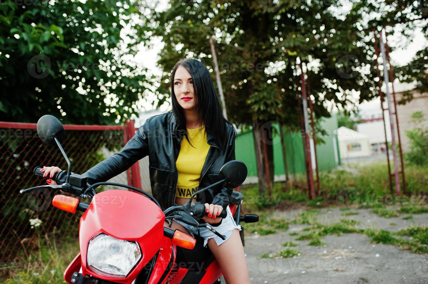 retrato de una mujer genial e impresionante con vestido y chaqueta de cuero negro sentada en una moto roja genial. foto