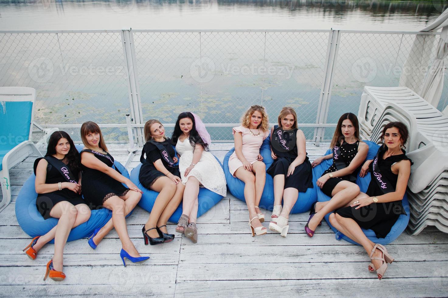 Group of 8 girls wear on black and 2 brides at hen party sitting on pillows at pier on beach side. photo