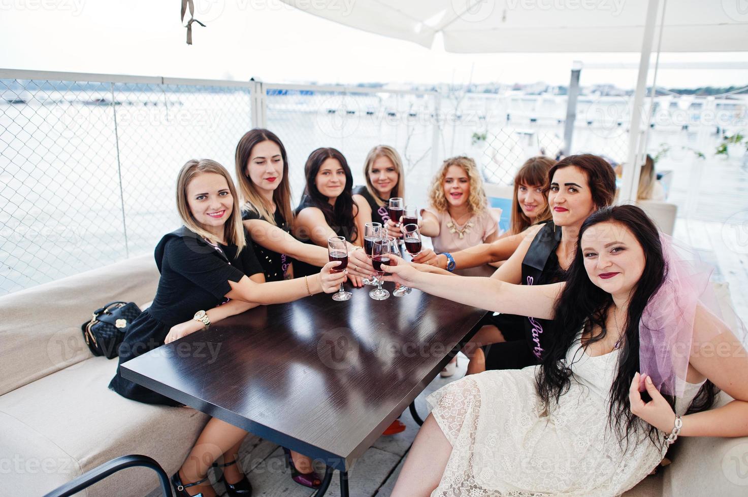 Group of 8 girls wear on black and 2 brides at hen party sitting at table and drinking rose champagne from glasses. photo