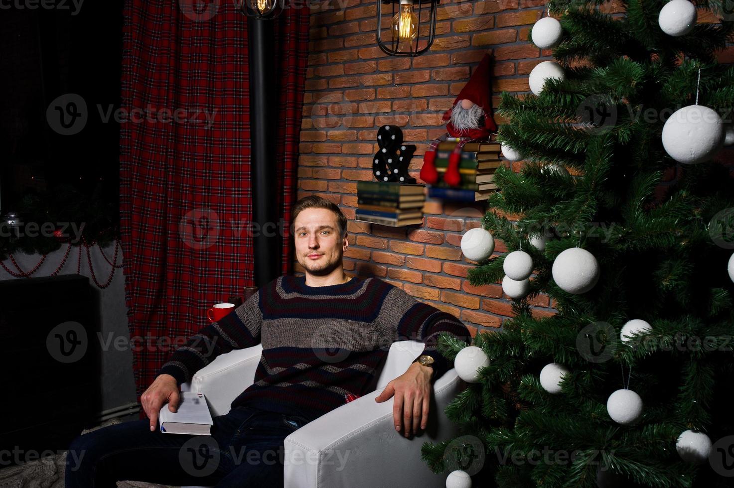 retrato de estudio de un hombre con un libro sentado en una silla contra un árbol de navidad con adornos. foto
