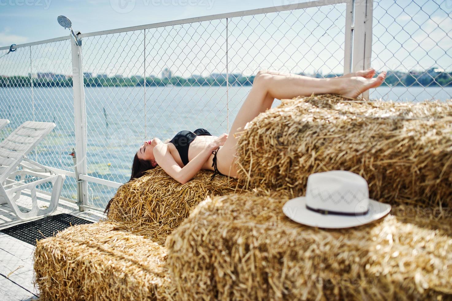 Portrait of a gorgeous girl in black bikini swimsuit posing on the hay bale with a hat by the lake. photo