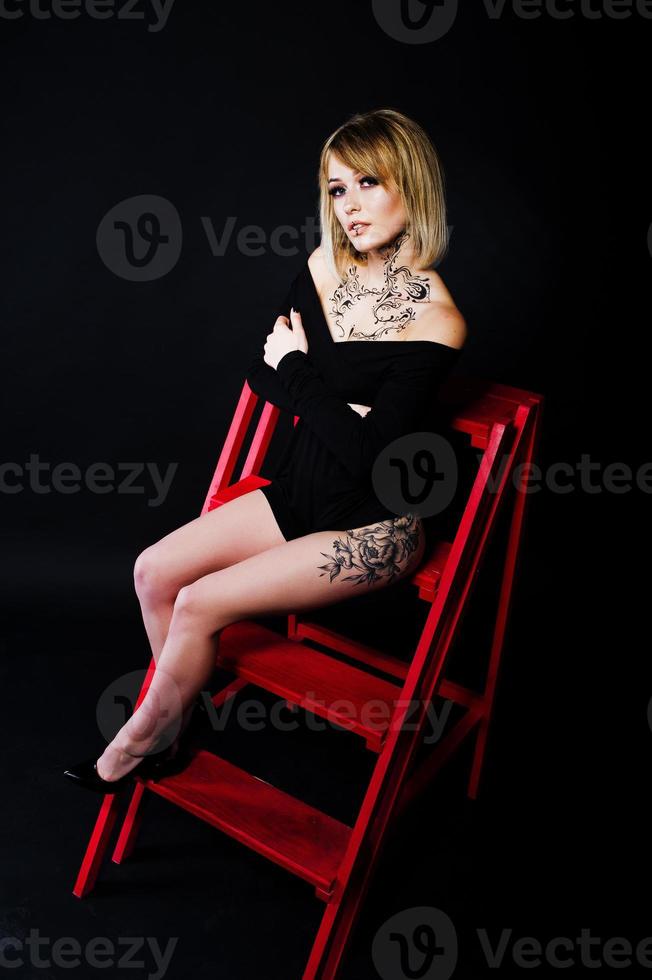 retrato de estudio de una chica rubia con maquillaje original en el cuello y tatuaje en el muslo, vestida con un vestido negro de fondo oscuro, sentada en una escalera roja. foto