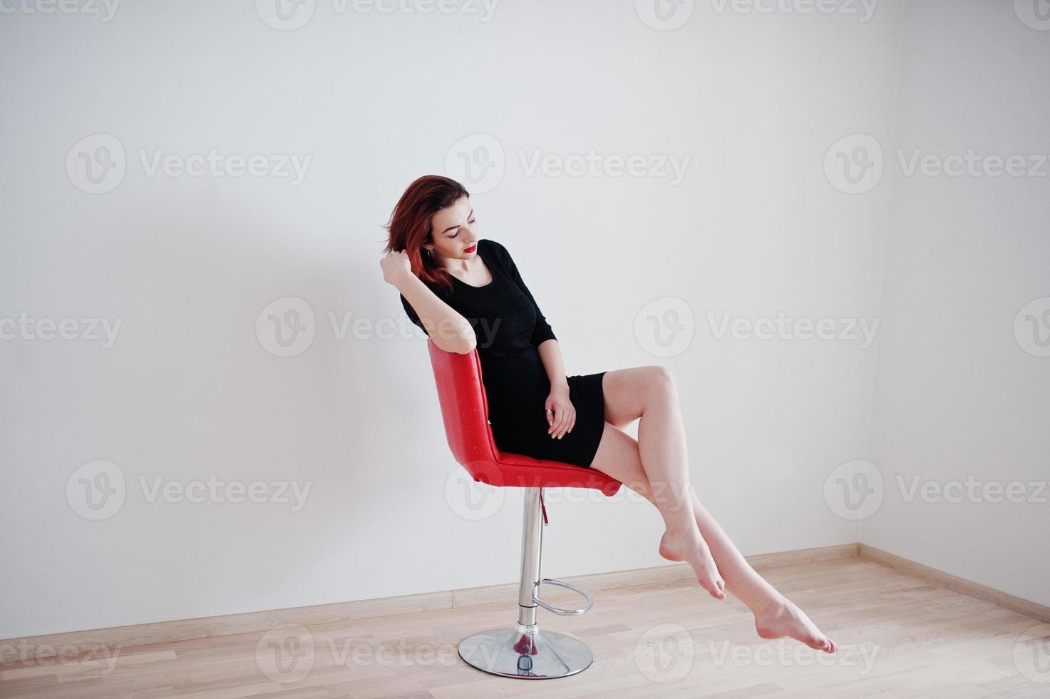 Red haired girl on black dress tunic sitting on red chair against white wall at empty room. photo