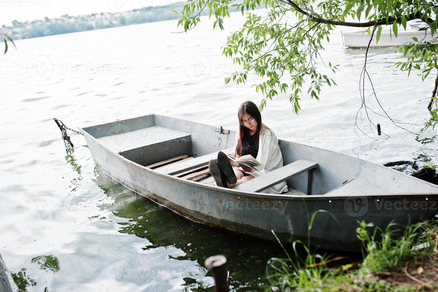 Portrait of an attractive woman wearing black polka dots dress, white shawl and glasses reading a book in a boat on a lake. photo