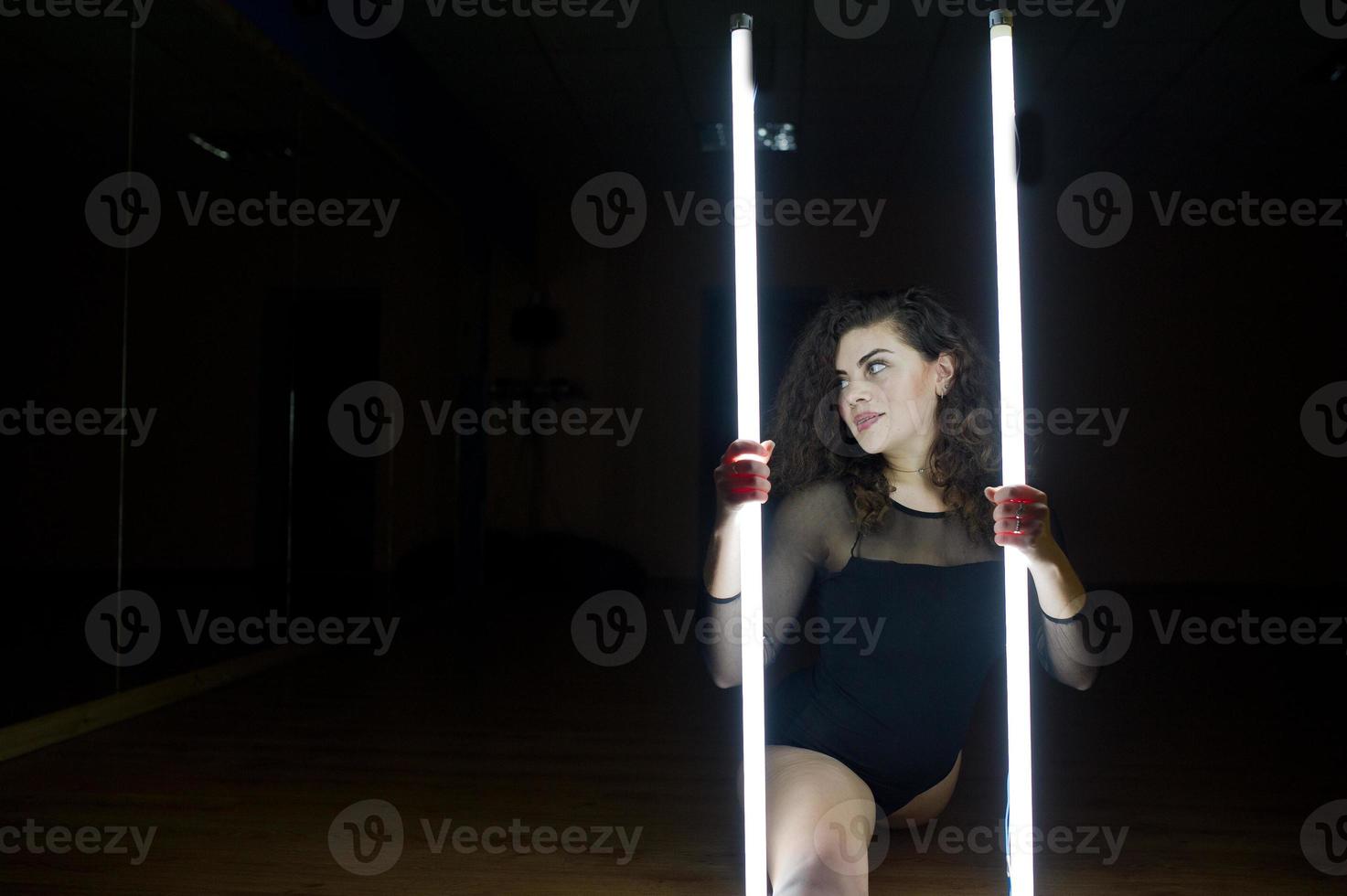 Curly brunette plus size model with two long led lamp at black room. photo