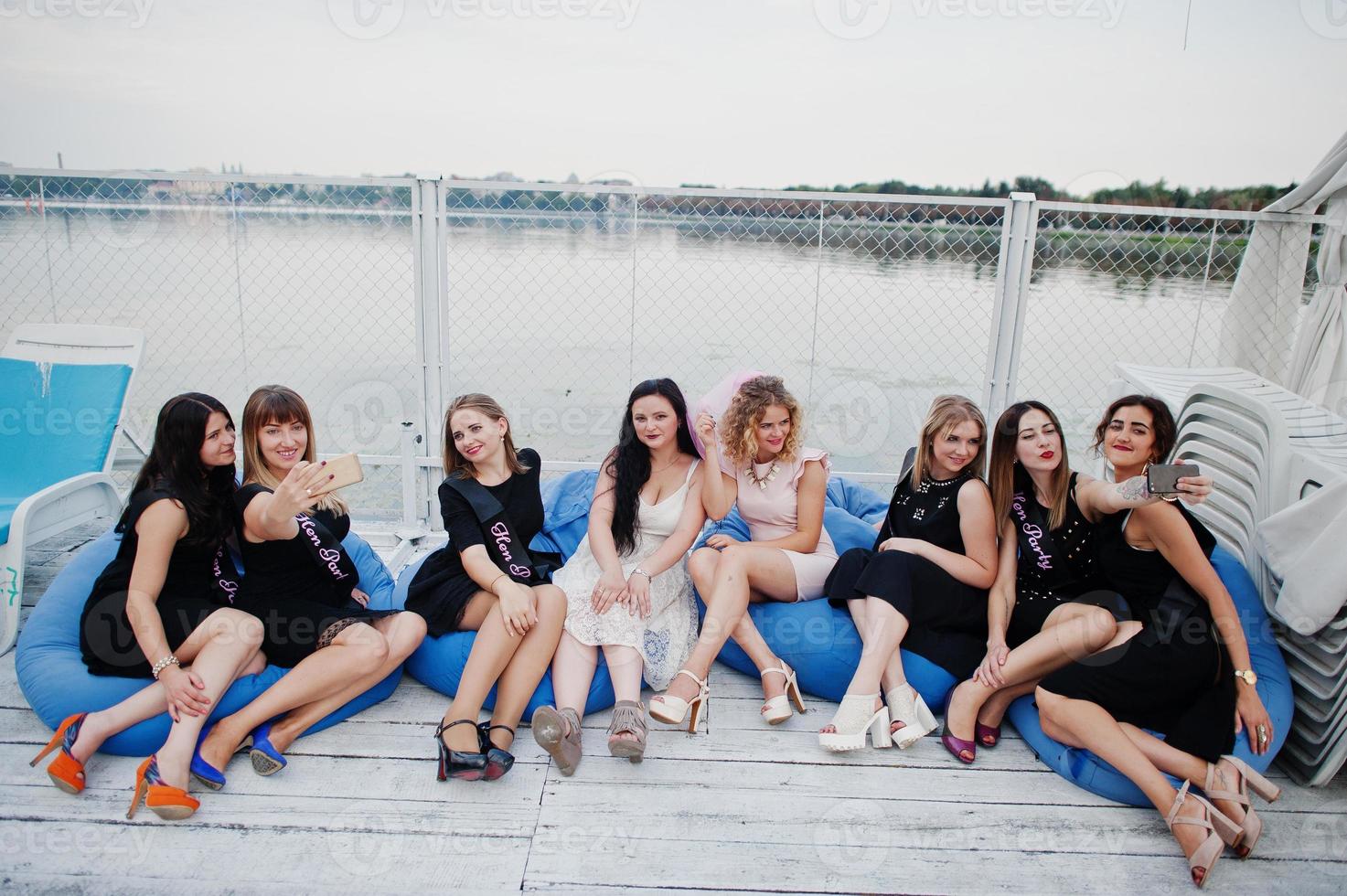 Group of 8 girls wear on black and 2 brides at hen party sitting on pillows and making selfie at pier on beach side. photo