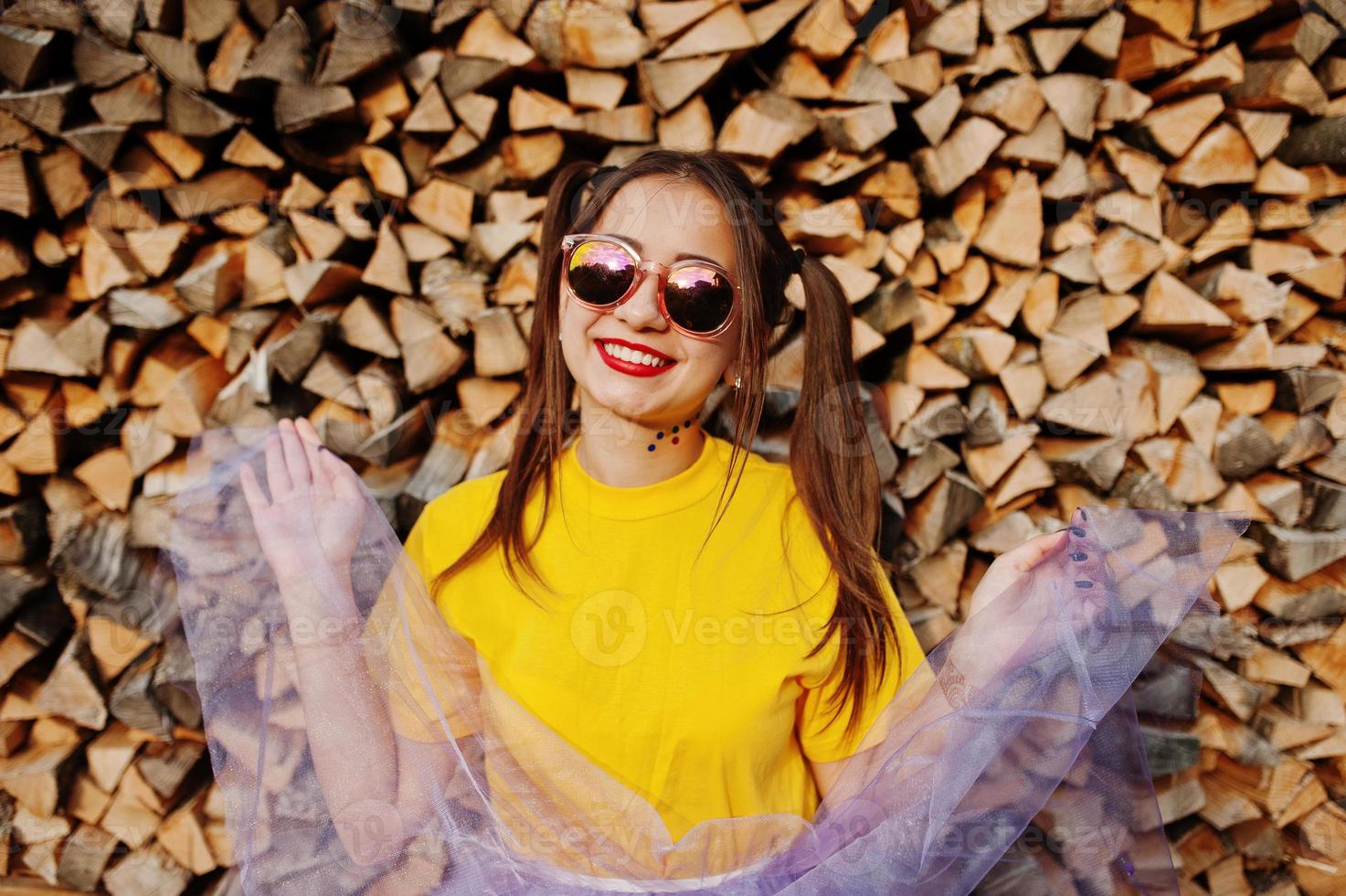 Young funny girl with bright make-up, tail hair wear on yellow shirt and sunglasses against wooden background. photo