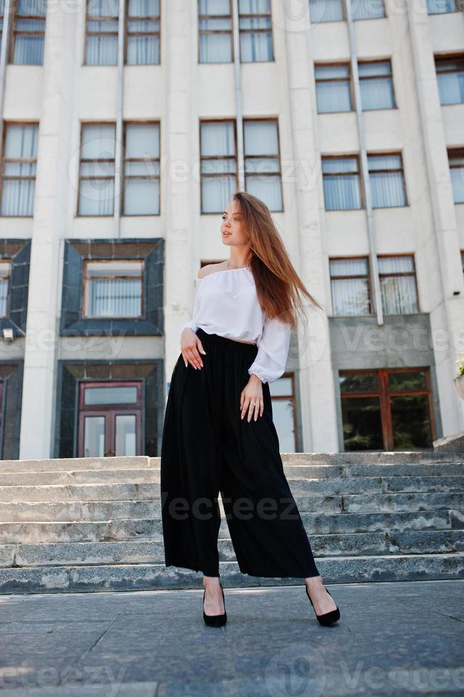 Portrait of a fabulous young successful woman in white blouse and broad black pants posing on the stairs with a huge white building on the background. photo