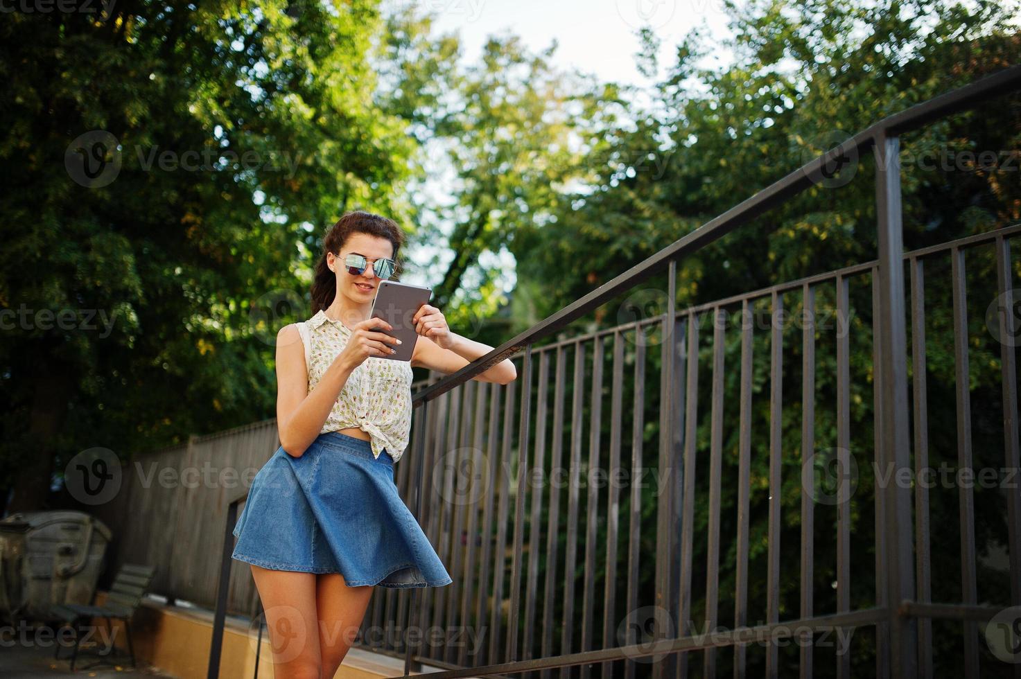 Curly stylish girl wear on blue jeans skirt, blouse with tablet on hands. photo