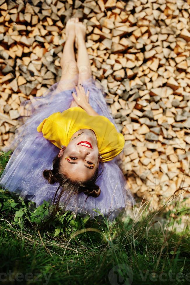 Young funny girl with bright make-up, like fairytale princess, wear on yellow shirt and violet skirt lying against wooden background. photo